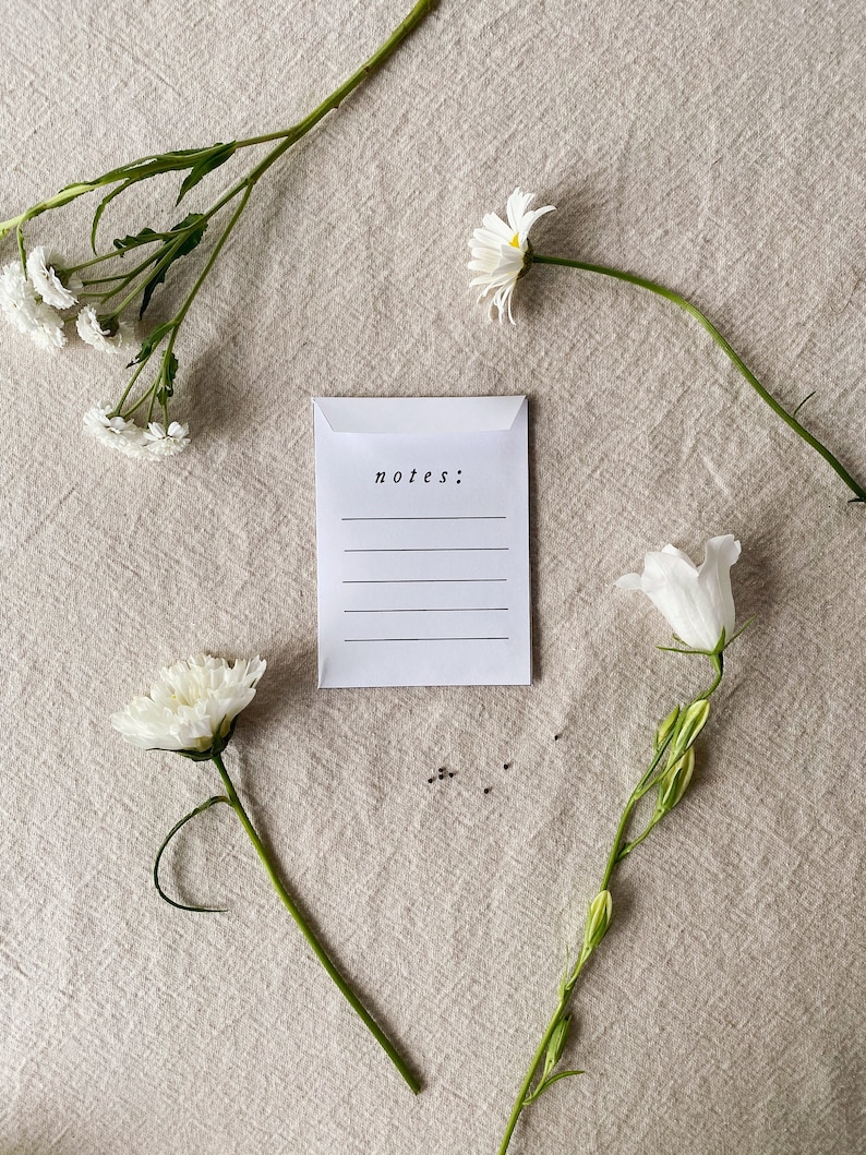 May include: A white envelope with the word "notes:" printed on it, with lines for writing. The envelope is surrounded by white flowers and green stems, arranged on a beige linen surface.