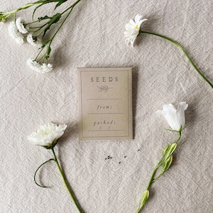 May include: A kraft paper seed packet with the word "SEEDS" and a botanical illustration, surrounded by white flowers and scattered seeds on a neutral linen surface. The packet has space for "from:" and "packed:" information.
