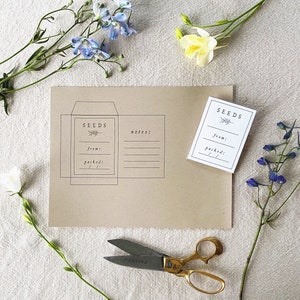 May include: A kraft paper seed packet and a small white seed label, both with the word "SEEDS" and space for notes. The packet and label are decorated with a sprig of leaves. Gold scissors and flowers are arranged around the packet.