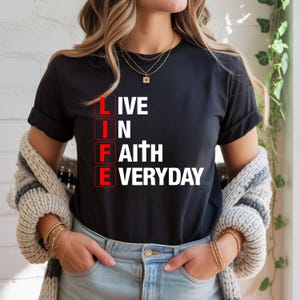 Può includere: T-shirt nero con le parole "LIVE LIFE IN FAITH EVERYDAY" in lettere bianche e rosse. La maglietta ha un girocollo e maniche corte. La modella indossa jeans blu e un maglione color crema.