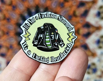 May include: A circular enamel pin with a pale yellow background featuring a black leather jacket over a green and white striped shirt. The text reads It's Not a Fashion Statement It's a Fucking Deathwish, accented by two yellow lightning bolt icons.