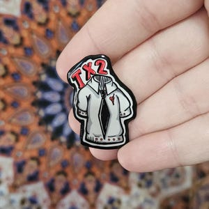 May include: A white shirt enamel pin with a black tie and a small red heart. The pin has the text "TX2" in red at the top. The pin is held in a hand.