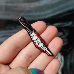 Puede incluir: Un pin de esmalte negro y blanco de un cuchillo ensangrentado con la palabra "INK" escrita en la hoja.