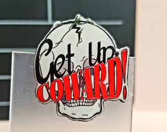 May include: A white skull-shaped pin with the words "Get Up COWARD!" in black and red. The pin has a cartoonish design with a cracked skull and a jagged lightning bolt. The pin is on a silver surface.