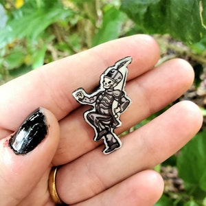 May include: A small, black and white enamel pin featuring a skeleton in a marching band uniform. The pin has a silver outline and is set against a background of green foliage. The skeleton is in a marching pose.
