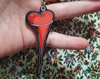 May include: A red heart-shaped keychain with a black outline and a silver keyring. The heart has a white design around the top and a long, pointed bottom. The keychain is held in a hand.