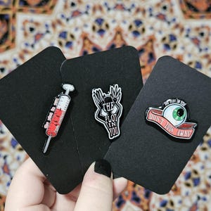 May include: Three enamel pins on black cards. One pin is a syringe with red liquid, another has a hand design with the text "THE PLOT IN YOU", and the last is an eye with the text "DON'T LOOK AWAY".