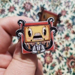 May include: A rectangular enamel pin featuring a cartoon character with a yellow, box-shaped head, wide eyes, and a gaping mouth. The character has brown liquid dripping from the chin, wears a white collared shirt, and is framed by black swirls.