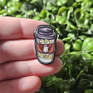 May include: A black and white enamel pin of a coffee cup with a ghost inside. The cup has a brown sleeve with the text "HOT MULLIGAN" in yellow letters.