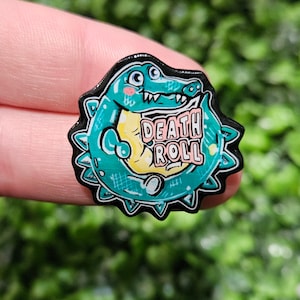 May include: A green alligator with a toothy grin and a pink and yellow "DEATH ROLL" text on its belly. The alligator is surrounded by black spikes.