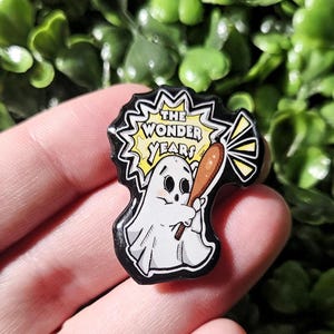 Puede incluir: Un pin de esmalte negro y blanco con un fantasma de dibujos animados sosteniendo un bate de béisbol. El fantasma tiene una expresión de sorpresa. El pin tiene un fondo amarillo con el texto "THE WONDER YEARS" en negro.