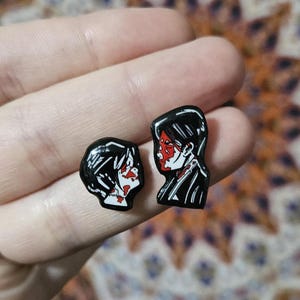 May include: Two black enamel pins featuring stylized portraits of a person with red accents. The pins depict a person in profile, with black hair and a white face, with red details. The pins are small and likely for clothing or accessories.