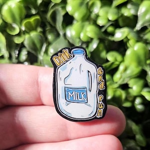 May include: A black and white enamel pin featuring a cartoon milk jug with the word "MILK" on the front. The pin has the words "Drink and Use" on the sides. The pin is held in a person's hand.