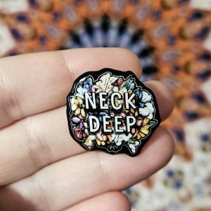 Op de afbeelding: Een zwarte emaille pin met een bloemenmotief en de tekst "NECK DEEP" in witte letters.