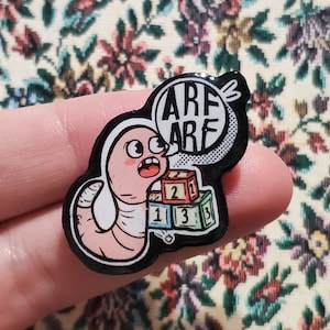 Könnte beinhalten: Ein Emaille-Pin im Cartoon-Stil mit einem rosa Wurm mit überraschtem Gesichtsausdruck. Der Wurm sagt "ARF ARF" in einer Sprechblase. Der Pin enthält auch drei farbige Bausteine mit den Zahlen 1, 2 und 3.