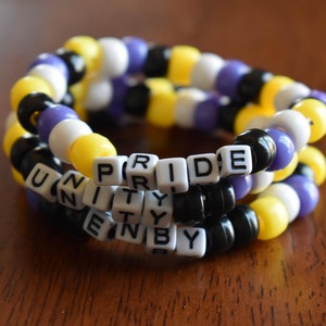 May include: Three beaded bracelets with the words "PRIDE", "UNITY", and "ENBY" spelled out in black letters on white beads. The bracelets are made with yellow, white, purple, and black beads.