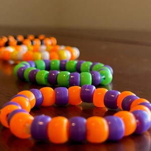 Op de afbeelding: Drie kralenarmbanden in oranje, paars, groen en zwart. De armbanden zijn gemaakt van kleine, ronde kralen die aan elkaar zijn geregen. De armbanden liggen op een bruin oppervlak.