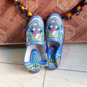 May include: A pair of blue embroidered slippers with colorful geometric patterns. The slippers are on a tiled floor.