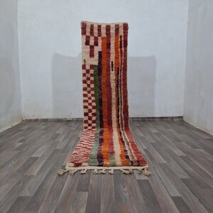 K&ouml;nnte beinhalten: Ein bunter Teppich mit geometrischem Muster und Fransen am unteren Rand. Der Teppich liegt auf einem Holzboden.