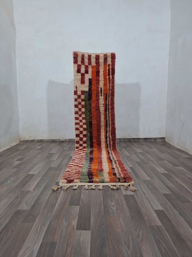 K&ouml;nnte beinhalten: Ein bunter Teppich mit geometrischem Muster und Fransen am unteren Rand. Der Teppich liegt auf einem Holzboden.