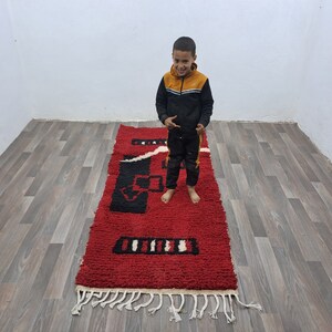 Puede incluir: Una alfombra rectangular roja con dise&ntilde;os geom&eacute;tricos en negro y blanco. La alfombra tiene una textura peluda y flecos blancos en los extremos. Est&aacute; colocada sobre un suelo de madera.