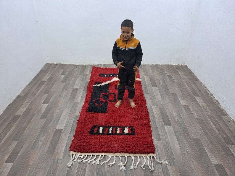 Puede incluir: Una alfombra rectangular roja con dise&ntilde;os geom&eacute;tricos en negro y blanco. La alfombra tiene una textura peluda y flecos blancos en los extremos. Est&aacute; colocada sobre un suelo de madera.