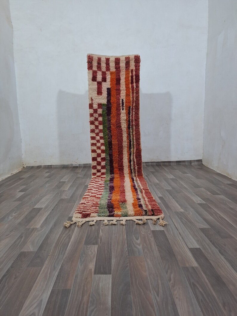 K&ouml;nnte beinhalten: Ein langer, rechteckiger Teppich mit einem geometrischen Muster in Rot-, Orange-, Gr&uuml;n- und Wei&szlig;t&ouml;nen. Der Teppich hat an beiden Enden Fransen.