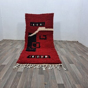 Puede incluir: Una alfombra bereber roja rectangular con un dise&ntilde;o geom&eacute;trico en negro y blanco roto. La alfombra tiene un pelo grueso y texturizado y una franja en el borde inferior. El dise&ntilde;o presenta formas rectangulares y cuadradas.