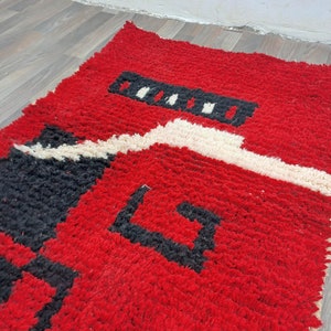 Puede incluir: Una alfombra rectangular tejida a mano con un fondo rojo vibrante. La alfombra presenta dise&ntilde;os geom&eacute;tricos en blanco y negro, creando un contraste llamativo. La textura parece afelpada y acogedora, lo que sugiere un revestimiento de suelo c&oacute;modo.