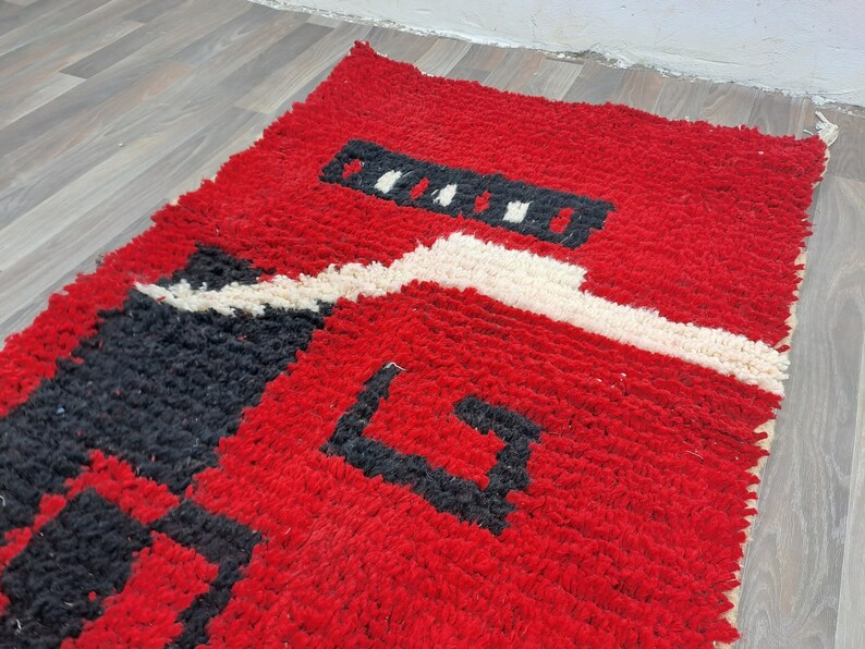 Puede incluir: Una alfombra rectangular tejida a mano con un fondo rojo vibrante. La alfombra presenta dise&ntilde;os geom&eacute;tricos en blanco y negro, creando un contraste llamativo. La textura parece afelpada y acogedora, lo que sugiere un revestimiento de suelo c&oacute;modo.