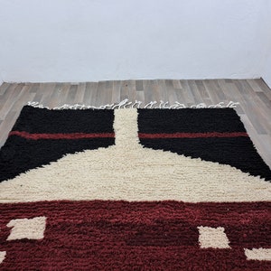 May include: A hand-woven rug with a geometric design in shades of red, black, and white. The rug has a thick, fluffy texture and is made with natural fibers.