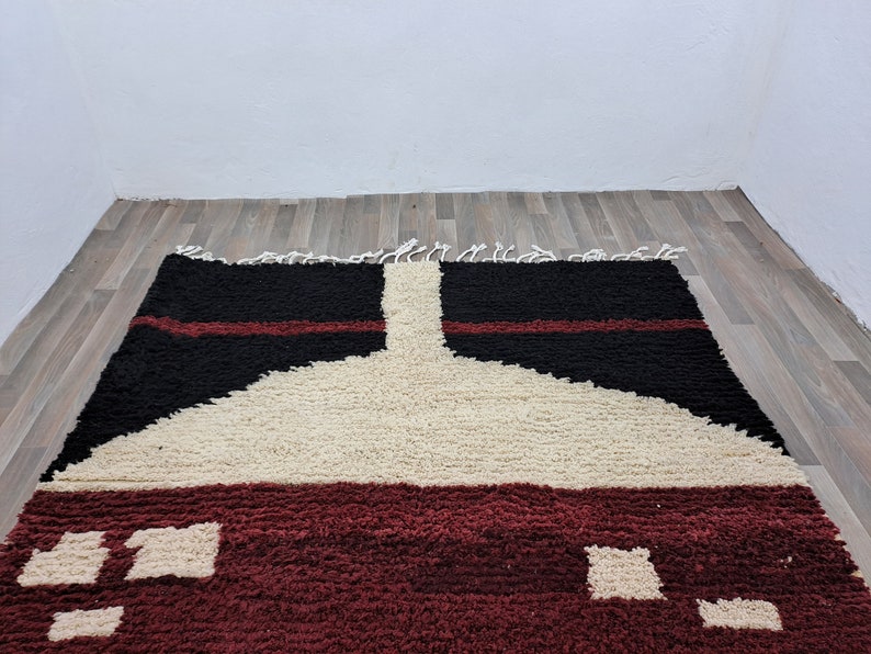 May include: A hand-woven rug with a geometric design in shades of red, black, and white. The rug has a thick, fluffy texture and is made with natural fibers.