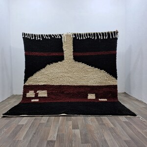 May include: A hand-woven rug with a geometric design in shades of black, white, and burgundy. The rug has a fringe border and is made of wool.