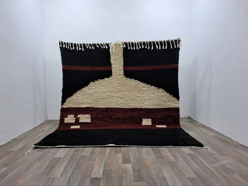 May include: A hand-woven rug with a geometric design in shades of black, white, and burgundy. The rug has a fringe border and is made of wool.