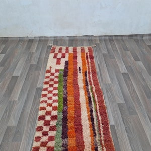 K&ouml;nnte beinhalten: Ein bunter Teppich mit geometrischem Muster mit roten, orangefarbenen, gr&uuml;nen und wei&szlig;en Streifen und karierten Mustern. Der Teppich liegt auf einem Holzboden.