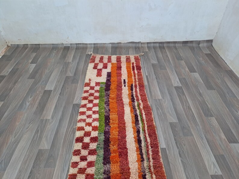 K&ouml;nnte beinhalten: Ein bunter Teppich mit geometrischem Muster mit roten, orangefarbenen, gr&uuml;nen und wei&szlig;en Streifen und karierten Mustern. Der Teppich liegt auf einem Holzboden.