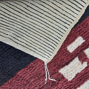 May include: Close-up of a multi-colored rug with a geometric pattern. The rug has black, white, red, and cream colored sections. The rug has a fringe on one edge.