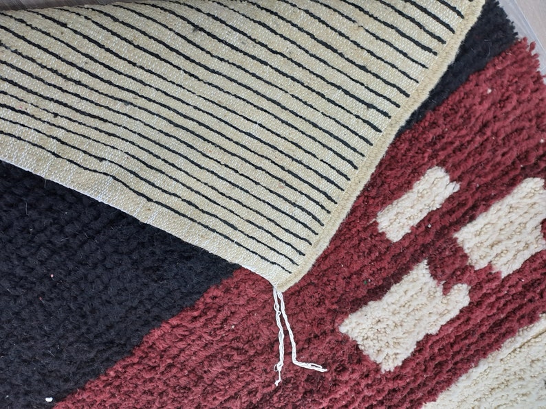 May include: Close-up of a multi-colored rug with a geometric pattern. The rug has black, white, red, and cream colored sections. The rug has a fringe on one edge.