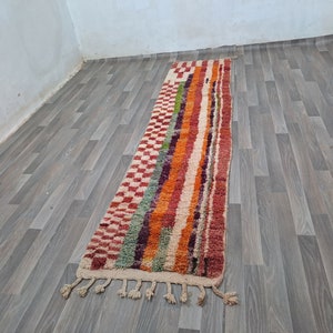 K&ouml;nnte beinhalten: Ein langer, bunter Teppich mit geometrischem Muster. Der Teppich hat an beiden Enden Fransen und ist aus Wolle. Der Teppich liegt auf einem Holzboden.