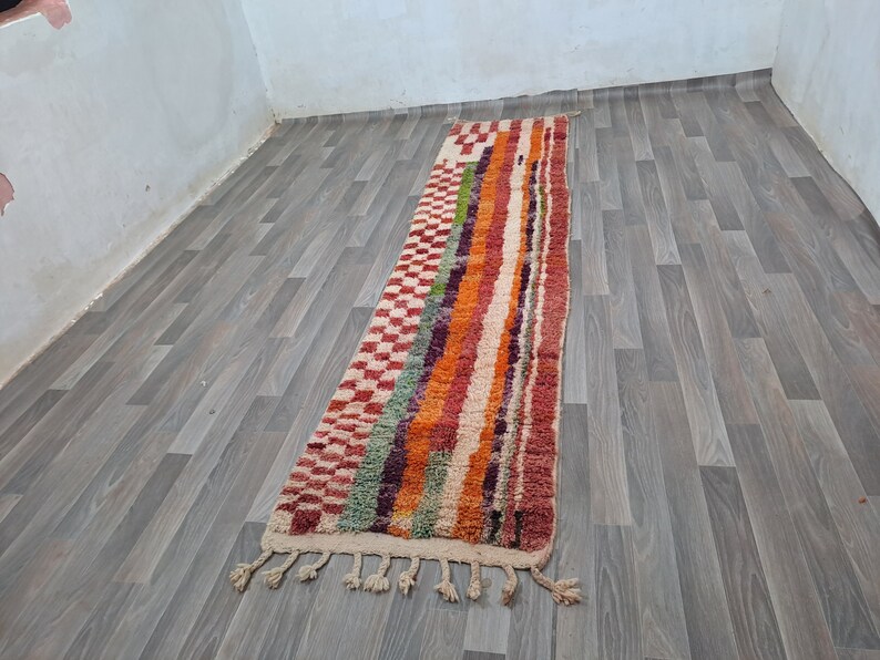 K&ouml;nnte beinhalten: Ein langer, bunter Teppich mit geometrischem Muster. Der Teppich hat an beiden Enden Fransen und ist aus Wolle. Der Teppich liegt auf einem Holzboden.