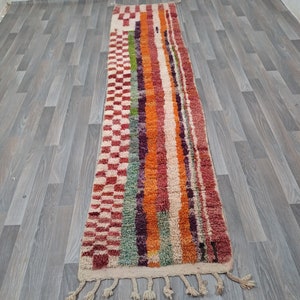 K&ouml;nnte beinhalten: Ein langer, rechteckiger Teppich mit einem bunten Streifenmuster. Der Teppich hat an beiden Enden Fransen und ist aus Wolle gefertigt. Der Teppich ist in verschiedenen Farben gehalten, darunter Rot, Orange, Gr&uuml;n, Lila und Wei&szlig;.