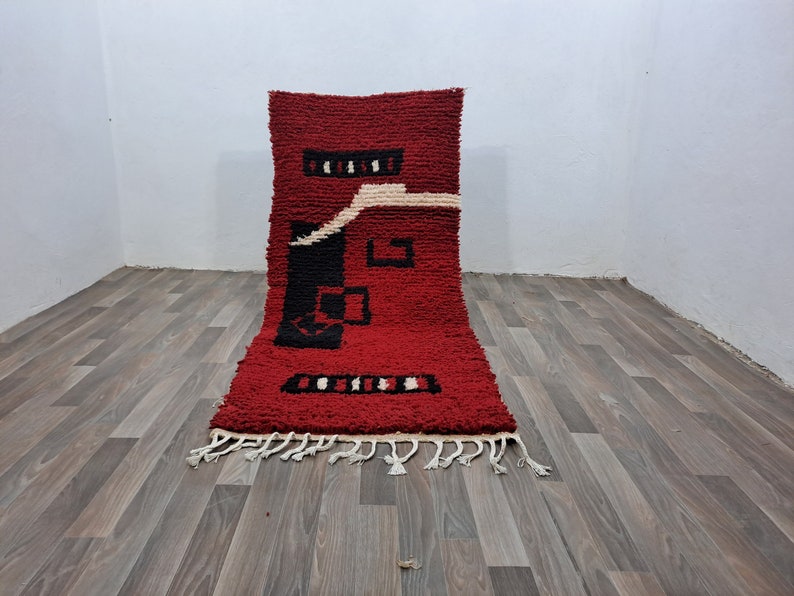 Puede incluir: Una alfombra rectangular roja con un dise&ntilde;o geom&eacute;trico en blanco y negro. La alfombra tiene un pelo grueso y texturizado y una franja en el borde inferior. El dise&ntilde;o incluye formas rectangulares y cuadradas.