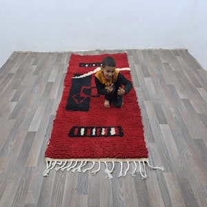 Puede incluir: Una alfombra rectangular roja con patrones geom&eacute;tricos en blanco y negro. La alfombra tiene un pelo grueso y texturizado y una franja a lo largo del borde inferior. La alfombra mide aproximadamente 152 cm de largo.