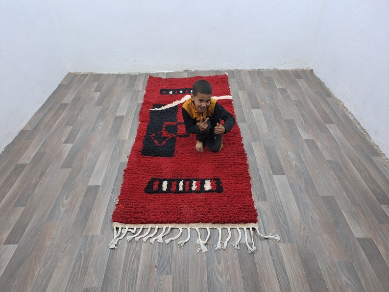 Puede incluir: Una alfombra rectangular roja con patrones geom&eacute;tricos en blanco y negro. La alfombra tiene un pelo grueso y texturizado y una franja a lo largo del borde inferior. La alfombra mide aproximadamente 152 cm de largo.