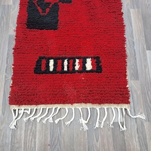 Puede incluir: Una vibrante alfombra roja con dise&ntilde;os geom&eacute;tricos en blanco y negro. La alfombra tiene un aspecto texturizado y peludo y un fleco de borlas blancas a lo largo de un borde. El dise&ntilde;o incluye una forma rectangular y l&iacute;neas verticales.