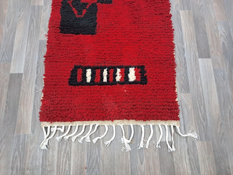 Puede incluir: Una vibrante alfombra roja con dise&ntilde;os geom&eacute;tricos en blanco y negro. La alfombra tiene un aspecto texturizado y peludo y un fleco de borlas blancas a lo largo de un borde. El dise&ntilde;o incluye una forma rectangular y l&iacute;neas verticales.