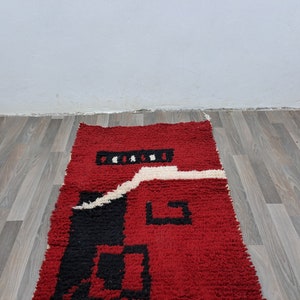 Puede incluir: Una alfombra rectangular roja con un dise&ntilde;o geom&eacute;trico. La alfombra presenta un campo rojo central con detalles en negro y blanco. El dise&ntilde;o incluye cuadrados negros y una l&iacute;nea blanca ondulada. La alfombra est&aacute; hecha de un material texturizado.