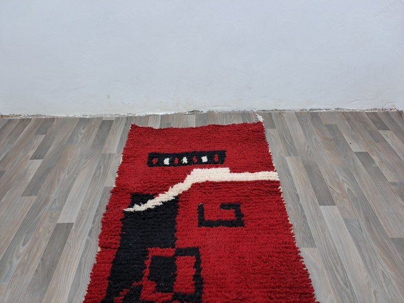 Puede incluir: Una alfombra rectangular roja con un dise&ntilde;o geom&eacute;trico. La alfombra presenta un campo rojo central con detalles en negro y blanco. El dise&ntilde;o incluye cuadrados negros y una l&iacute;nea blanca ondulada. La alfombra est&aacute; hecha de un material texturizado.