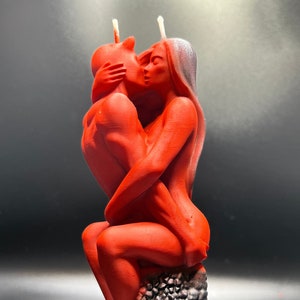 Puede incluir: Una escultura de vela roja de una pareja besándose. La pareja está sentada sobre una base texturizada de color gris.