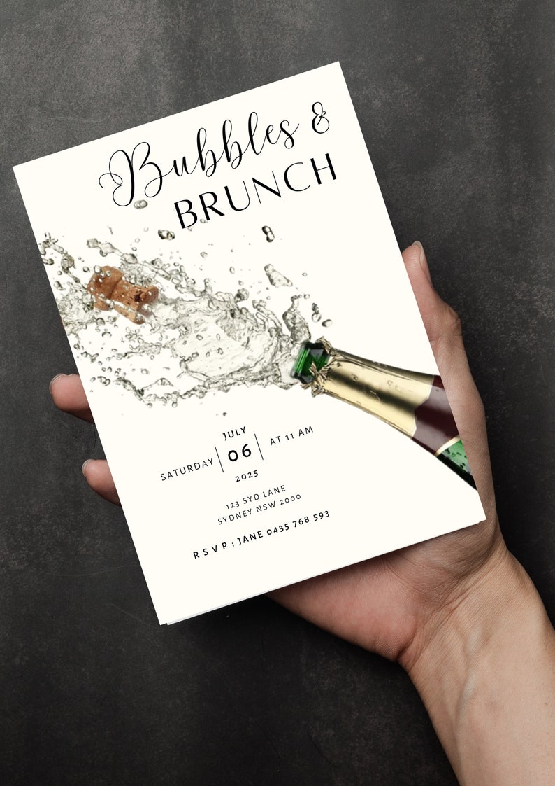 May include: A white invitation card with black text for a Bubbles & Brunch event. The invitation features a graphic of a champagne bottle with a cork popping out and a spray of bubbles. The text on the invitation includes the date, time, and location of the event. RSVP information is also included.